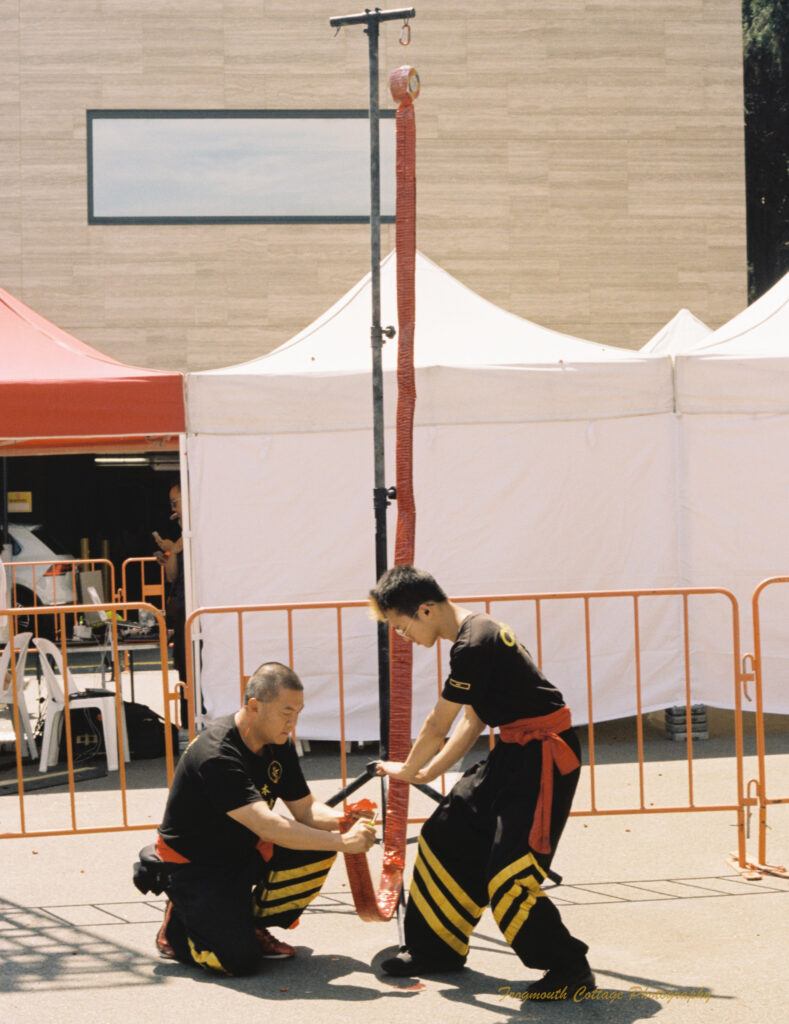 Photo of two chinese men, dressed in black costumes with red and gold trim, lighting the end of a red firecracker ribbon that is hanging from a tall pole.