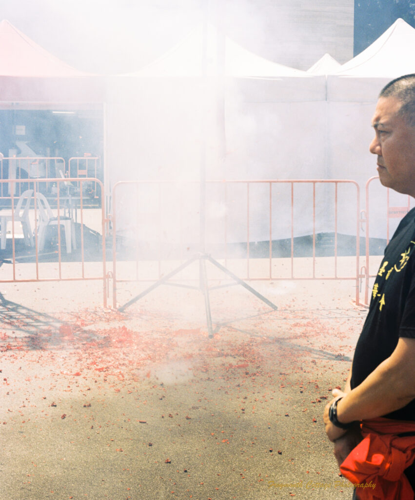 Photo of a chinese firecracker ribbon hanging on a pole going off. The photo is filled with smoke, there are bits of red paper beneath and around the pole the cracker is on. A chinese man stands to the side with his hands claspsed in front, supervising.