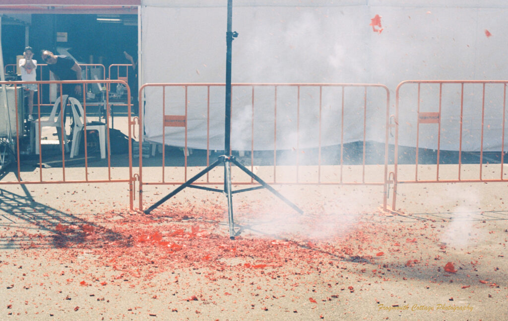 Photo of the area beneath a chinese firecracker pole. There is smoke where crackers have fallen and lots of red paper on the ground, and some in the air. In the background a young boy and man are watching, the boy has his hands over his ears. The man is bending down for a better view.