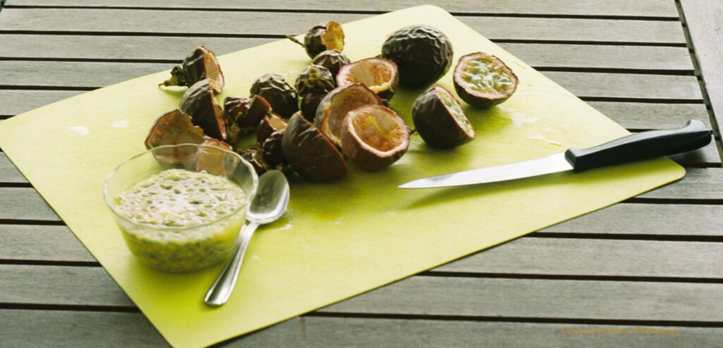 Close up color photograph of passionfruits sliced in half with a bowl of pulp, knife and teaspoon on a yellow chopping board.