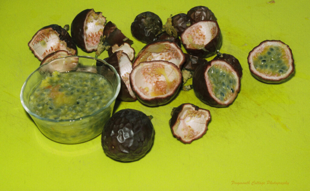 Close up color photograph of passionfruits sliced in half with a bowl of pulp, knife and teaspoon on a yellow chopping board.