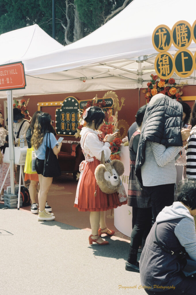 Photo of a line of people in front of a chinese new year stall. The stall has a white tent roof and red walls. A woman dressed in a red skirt and shoes is carrying a handbag that is a teddy bear face.