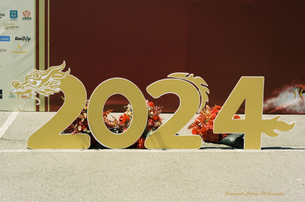 A gold coloured "2024" sign sitting on the ground in front of a red wall. The first "2" has the head of a drag on it, the "4" has a tail.