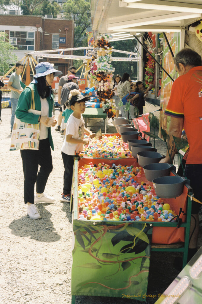 Photo looking down a line of sideshow stalls. A boy and his mother are looking at a game consisting of hundreds of coloured balls floating in trays of water.