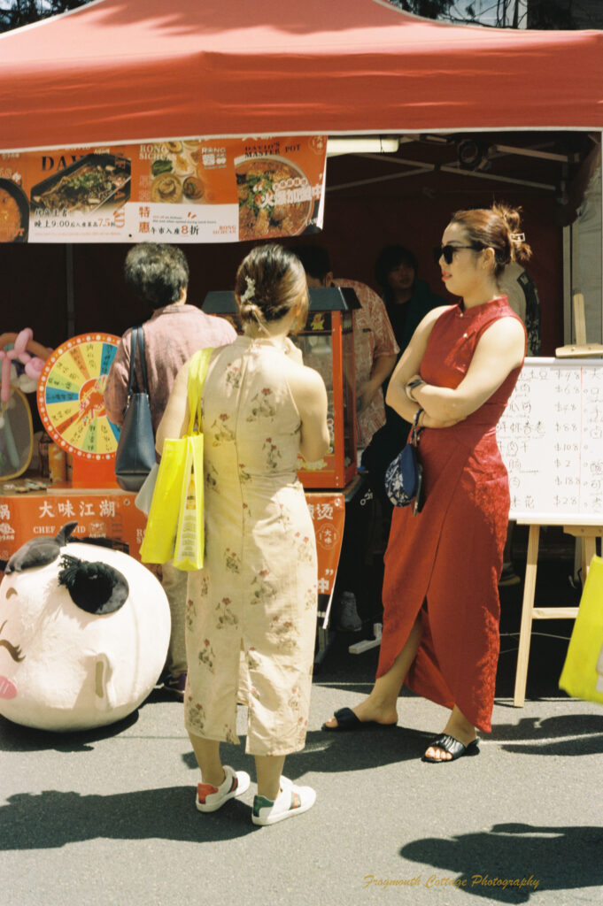 Photo of a woman in red dress and a woman in a cream dress with red flowers standing in the sun in front of a chinese food stall.