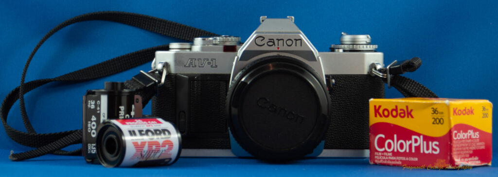 Photograph of a Canon AV-1 SLR camera with a strap and three rolls of film.