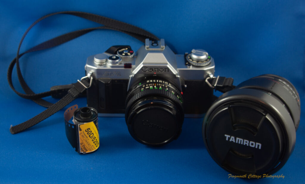 Photograph of a Canon AV-1 SLR camera with a strap, a roll of film and a Tamron zoom lens next to it.