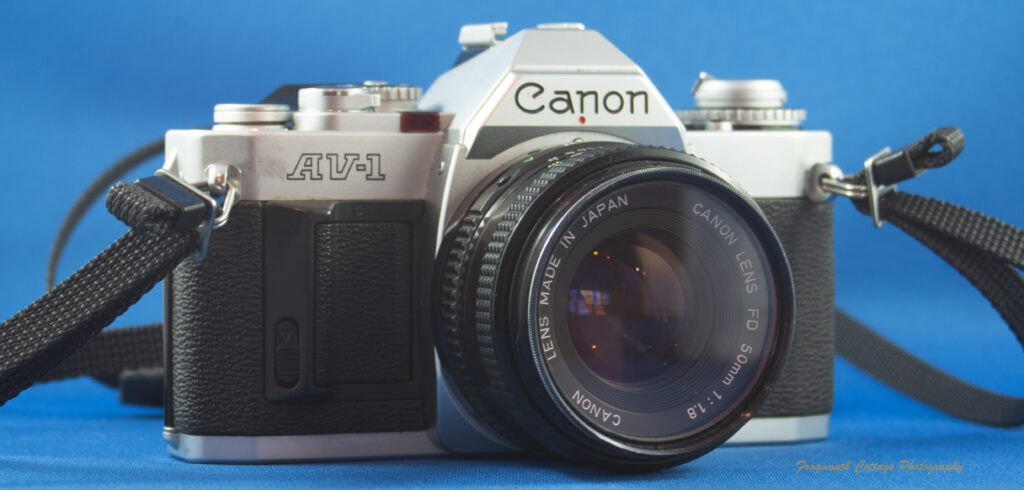 Photograph of a Canon AV-1 SLR camera with a strap. The lens cover has been removed and the front of the FD 50mm lens is visible.