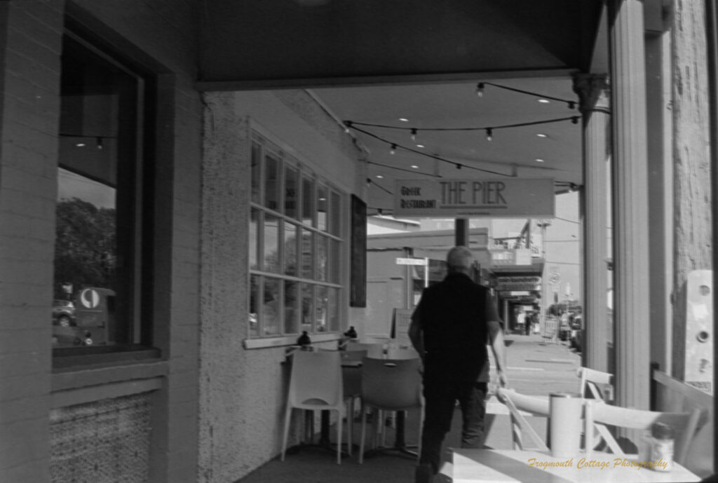 Black and white photo of a man walking away from the camera past tables on a footpath outside a restaturant. There are lights hanging from the verandah over the tables. The sign above the man reads "The Pier Greek Restaurant".