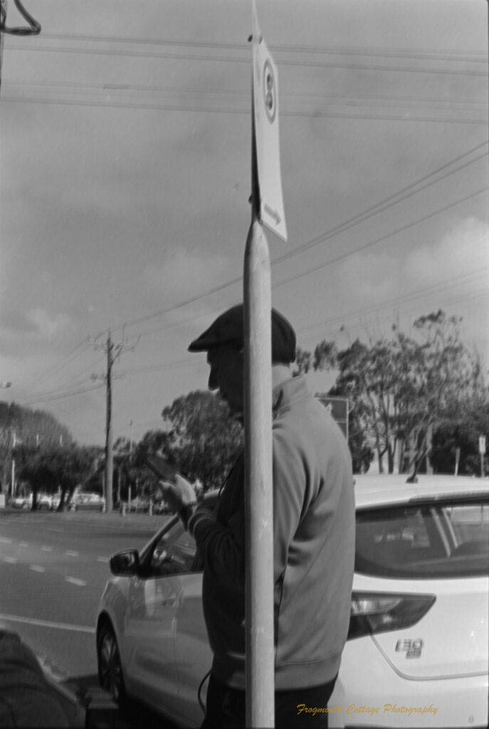 Black and white photo of a man leaning against a sign post. The man is looking at his phone. The sign syas "no standing".