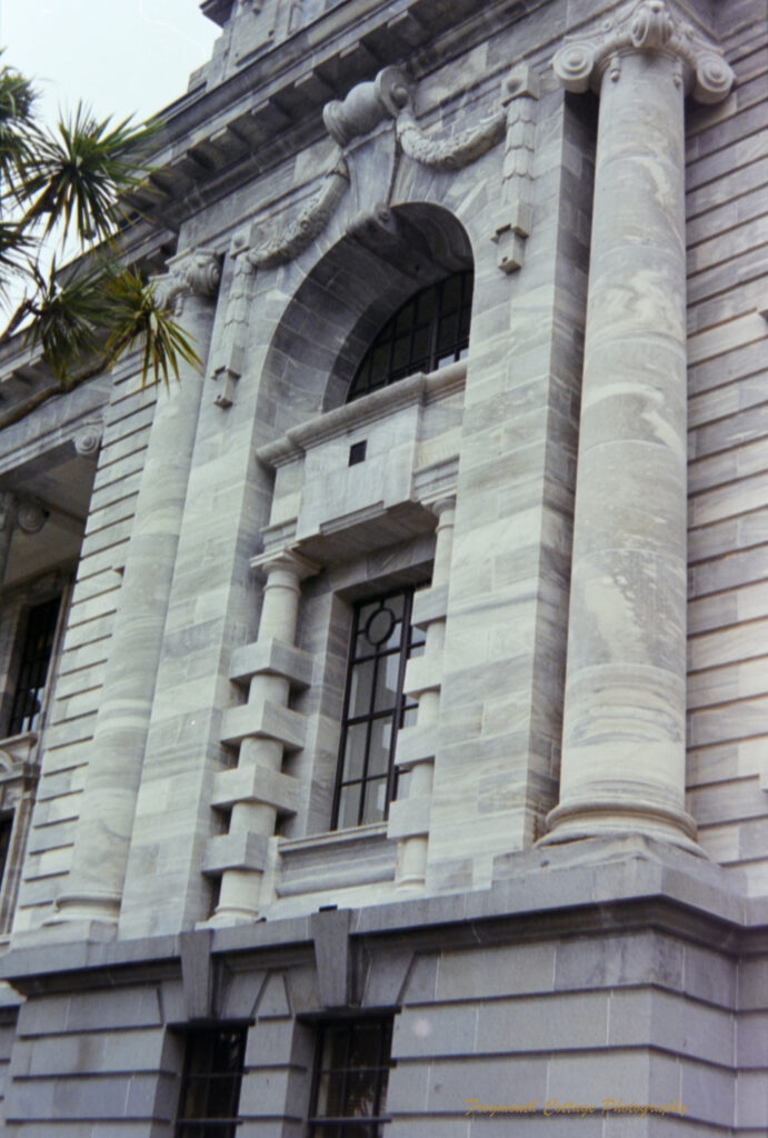 Photograph of the outside of a stone building looking up along columns that are either side of a large arched window. The columns are smooth grey stone in front of horizontal grey bricks.