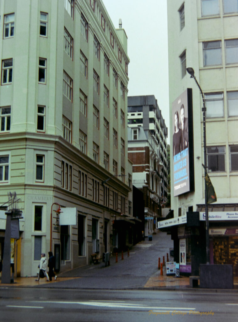 Photograph looking up an alley way of a larger street. There are tall cream coloured buildings eitherside of the alley and two people have just crossed in front of it. There is a large advertising billboard on one of the buildings. The sky is grey and it has been raining.