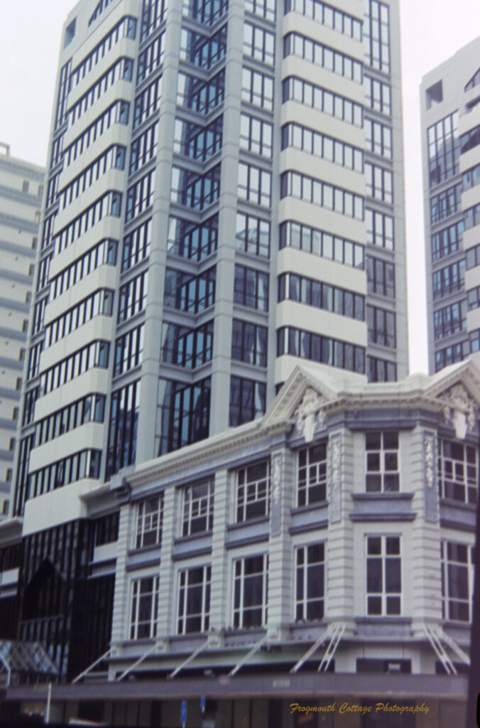 Photograph of a tall building that is predominatly black framed glass with white horizontal panels. It is next to a small whote stone building with white framed windows.