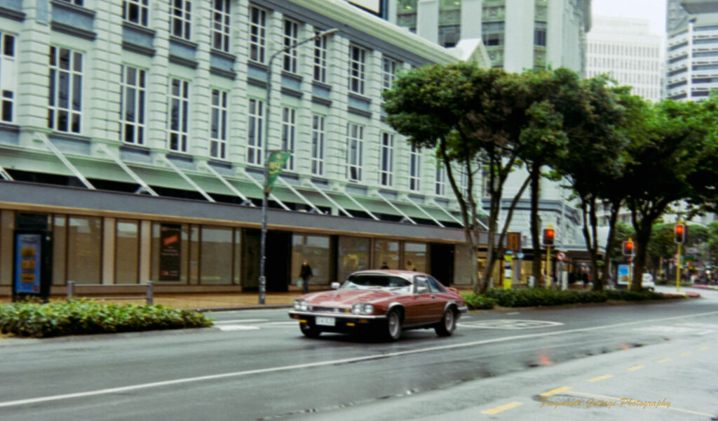 A photograph of a quiet main street with a single red classic car driving along it. Alongside the street is a large white stone building. It is a grey rainy day. The traffic lights at the intersection behind the car are glowing red.