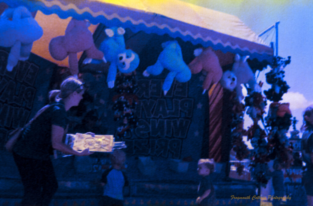 A photo of a mother and two children standing in front of a carnival game with large toys hanging from the top. The colour has broken up on the film and the greens and blues contains yellow patches.