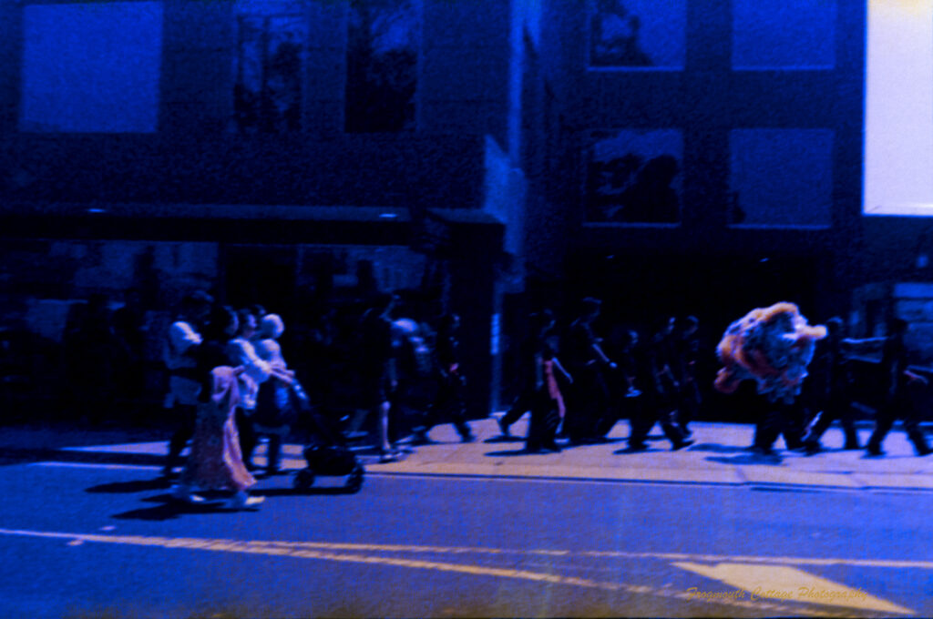 Photo of people walking along a street following a person who is carrying a chinese lion head after a performance. The image is dark and graining, but the lions head is colourful.