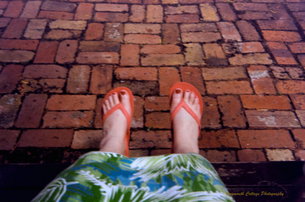 Colour photo of a pair of feet in red thongs hanging out over a paved brick path. The toes are painted red and the bottom of a green flowery dress can be seen above the feet.