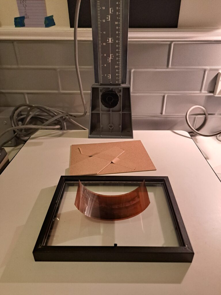 A photo frame lying glass side down, with the back removed and placed behind it. A negative has been placed on the glass and is curling upwards.