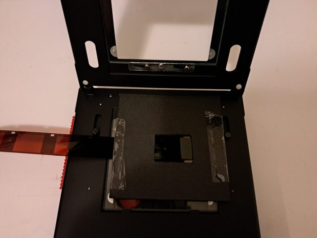 Photograph of a negative carrier with 110 film inserted and a cardboard mask over the top to create a frame around the smaller negatives.