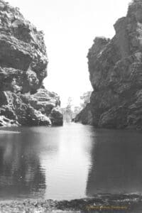 Black and white photograph of a large watering hole between two large rocky outcrops.