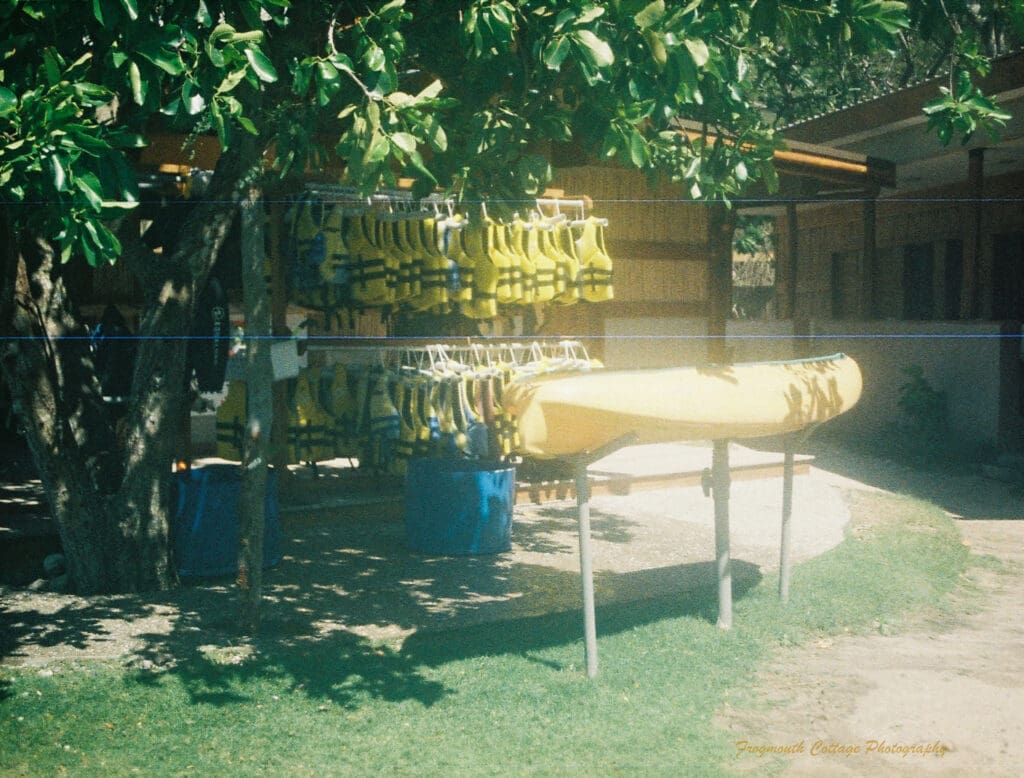 Photograph of a yellow canoe on a high stand in front of two rows of hanging yellow life jackets. There are thatched buildings in the background and lush green foliage from a large tree over the top.