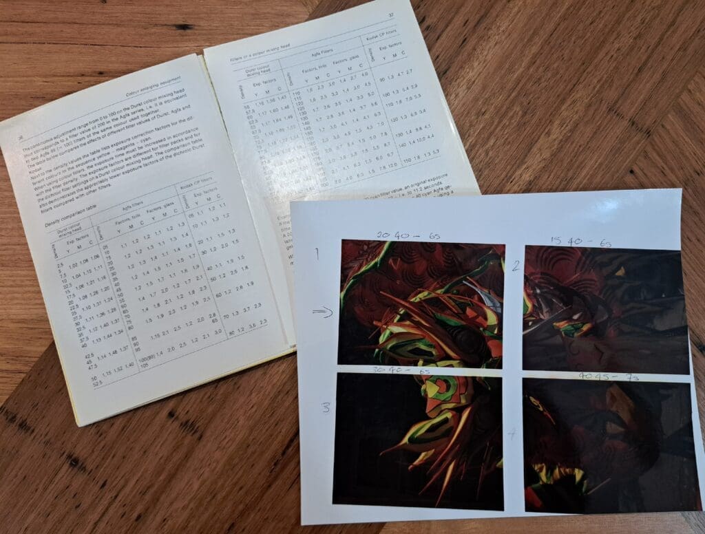 Photograph of a book open at a page with tables of densities for the Durst colour mixing head and Agfa and Kodak filters. Lying across it is a piece of paper with 4 images printed on it, labelled 1,2,3 and 4. There are notations of filter factors and exposure times next to each print. An arrow is pointing at the image labelled 1.
