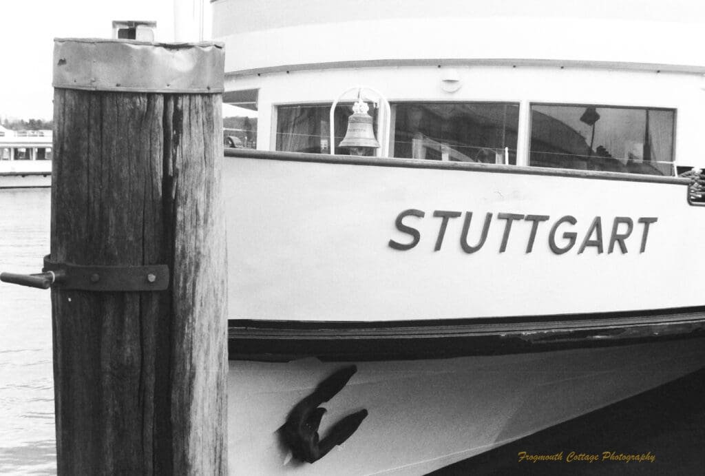 Black and white photo of the side of a wooden boat with the name "Stuttgart on the side of it. A wooden pier is in front of the boat.