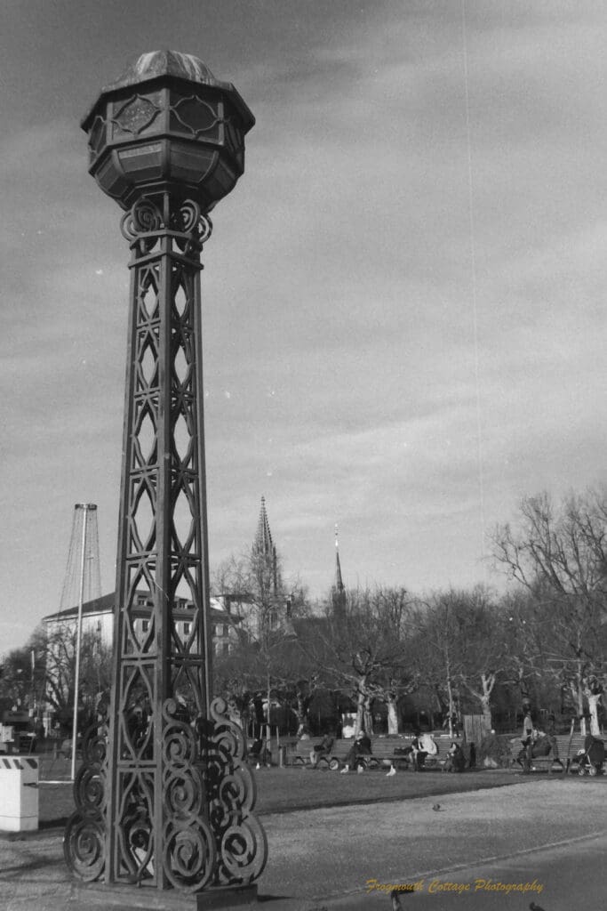 Black and white photograph of a park with a tall iron sculpture in the foreground.