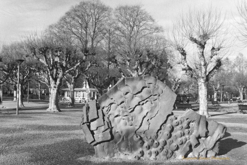 Black and white photograph of a park with bare trees and and a large stone sculpture in the foreground.