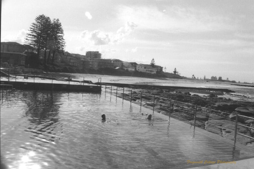 Black and white photograph of a swimming pool built into the rocks beside the ocean. There are two people in the pool and houses along the coast line in the background.