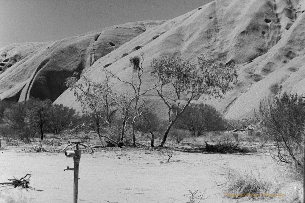 Black and white photo of the side of Uluru with a water tap in the foreground.
