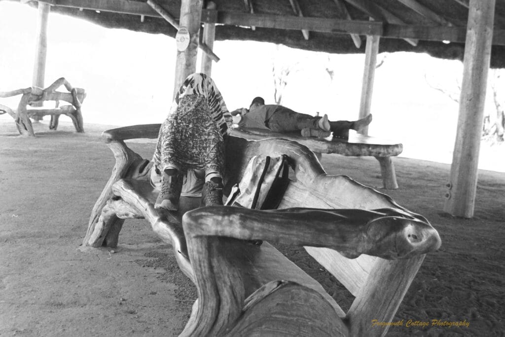 Black and white photo of two people resting under a covered area. One is lying on a table, the other is sitting on a bench seat with a sarong covering their head. The furniture is made of highly polished timber in it's natural form.