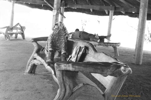 Black and white photo of two people resting under a covered area. One is lying on a table, the other is sitting on a bench seat with a sarong covering their head. The furniture is made of highly polished timber in it's natural form.