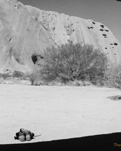 Uluru Mala Carpark Black and white photo taken in the shadow of a sheltered area. The roof has created a curved shadow on the ground. Two pairs of boots are sitting on the ground in front. The side of Uluru is in the background.