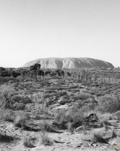 Uluru Black and white photo of a dark scrubby landscape with a large, long rock in the background.