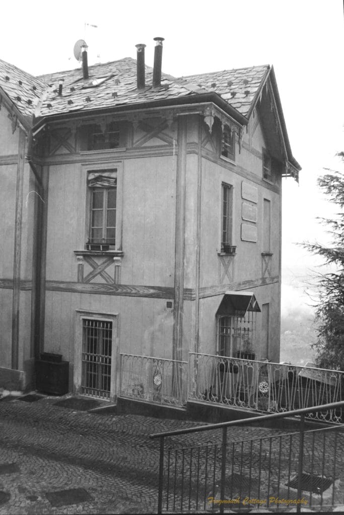 Black and white photograph of a two story house on hillside with a paved path and stair case rail in the foreground.